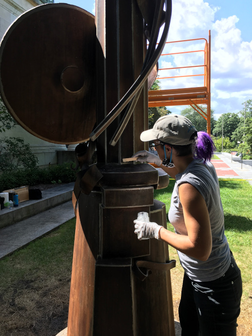 Photograph of a woman with a hat and short purple ponytail working outdoors. She is wearing gloves and applying a chemical gel to a tall metal sculpture.