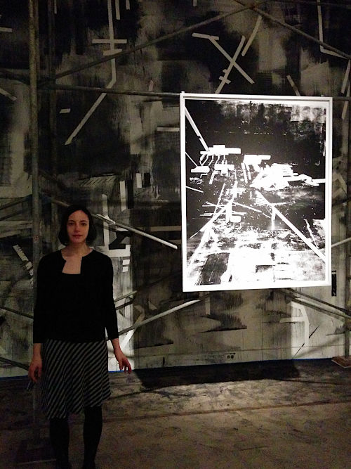 Photograph of a pale olive woman with chin-length dark hair and black clothing standing in an art installation with metal scaffolding and printed and painted black and white abstract forms.